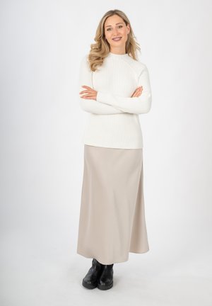 Witte geribbelde gebreide trui, beige satijnen maxi rok en zwarte chunky enkelboots. Model staat met gekruiste armen tegen een effen achtergrond.
