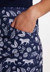 Námořnická látka s bílými květinovými a leopardími vzory. Pohled zblízka na otevřenou kapsu po straně, ukazující složité detaily a texturu.