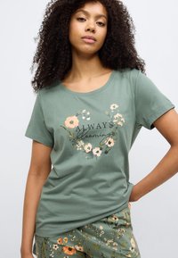 Πράσινο βαμβακερό μπλουζάκι με κοντά μανίκια, διακοσμημένο με λουλουδάτο μοτίβο και το κείμενο "Πάντα ανθίζω" σε κυκλικό σχέδιο. Λείο υλικό, άνετη εφαρμογή.