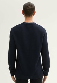Pull en maille bleu marine avec un motif texturé, col rond, manches longues, et poignets et ourlet côtes. Vue de dos, aucun détail supplémentaire visible.