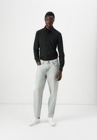 Camisa negra de botones, jeans azul claro y zapatillas blancas. El modelo está de pie sobre un fondo blanco sencillo, mostrando un corte relajado y un diseño simple.