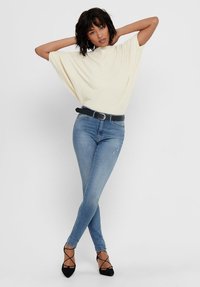 Cream-colored, loose-knit top with wide sleeves, paired with fitted light blue jeans and black strappy heels. Body positioned with arms raised.