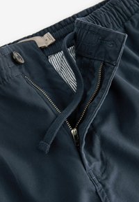 Pantalon bleu marine avec taille élastique, cordon de serrage, bouton et fermeture éclair métallique partiellement ouverte révélant une doublure intérieure rayée.