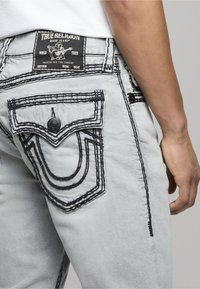 Vaqueros de mezclilla gris claro con costuras negras, un diseño de bolsillo único y una etiqueta con la marca "True Religion" en la cintura.