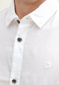 Weißes Leinenhemd mit Button-Down-Kragen, ausgestattet mit grauen Knöpfen und einem dezenten, in Weiß eingestickten Logo-Detail auf der Brust.