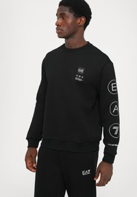Black sweatshirt with ribbed crew neck, featuring front and sleeve logos in white. The fabric appears soft and smooth.