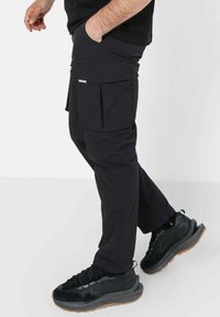 Svarte cargo bukser med slim fit, featuring store sidelommer, en glatt tekstur og et minimalistisk design. Kombinert med svarte joggesko.