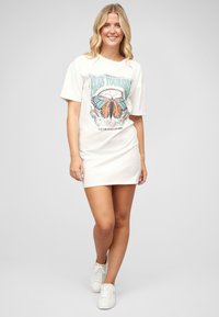 Biała sukienka t-shirtowa z krótkim rękawem, z kolorową grafiką motyla i tekstem. Miękka tkanina, luźny krój, noszona z białymi trampkami.