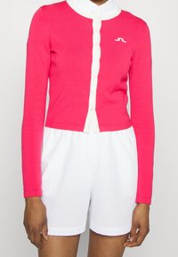 Ljusrosa långärmad cardigan med vit knappknäppning, som matchas med vita strukturerade shorts. Cardiganen har en liten logotyp på bröstet.