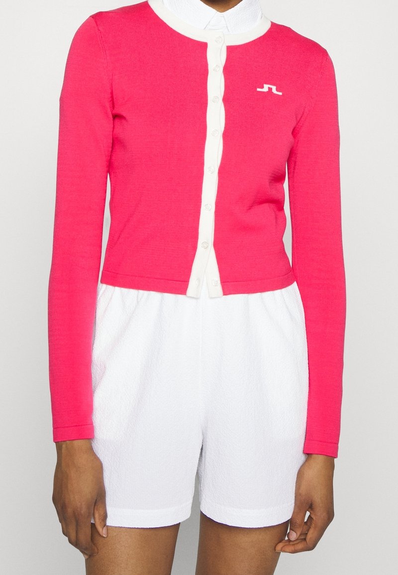 Ljusrosa långärmad cardigan med vit knappknäppning, som matchas med vita strukturerade shorts. Cardiganen har en liten logotyp på bröstet.