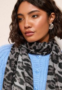 Szalik w cętkę lamparta w odcieniach szaro-czarnych, zarzucony na szyję. W tle można zobaczyć jasną, neutralną ścianę oraz fakturowany niebieski sweter.