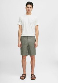 Hvit bomull t-skjorte med liten tekstlogo, kombinert med olivengrønne linshorts. Modellen har på seg brune sandaler. Ensfarget bakgrunn.