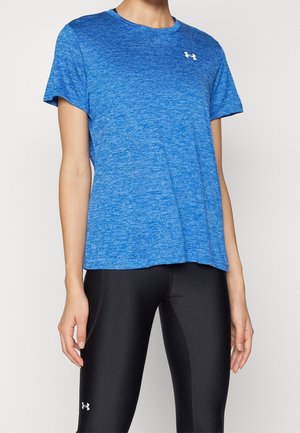 Osoba nosząca niebieską sportową koszulkę z krótkim rękawem i czarne dopasowane legginsy z logo Under Armour na koszulce i legginsach.