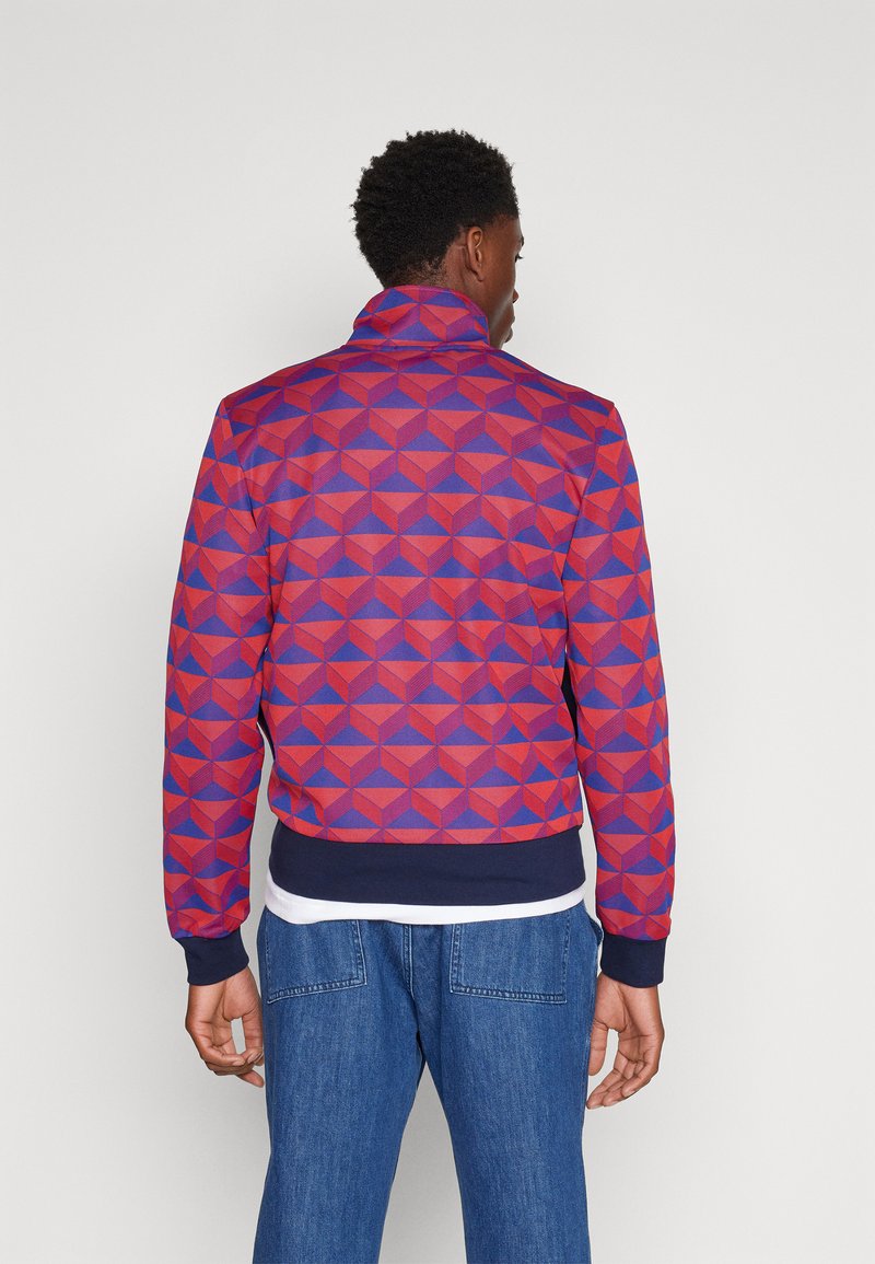 Homme portant une veste à motifs géométriques rouges et violets avec des poignets et une ceinture navy, associé à un jean bleu, de dos contre un fond uni.