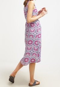 Kvinna i ärmlös, midiklänning med rosa och lila cirkulära mönster, iklädd svarta sandaletter, som går mot en enkel vit bakgrund.