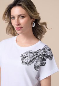Witte t-shirt met een zwart pailletten strikontwerp op één schouder, korte mouwen, ronde hals en een gladde stoftextuur.