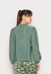 Blusa de seda verde com um decote franzido, mangas longas bufantes e uma textura suave, combinada com calças de padrão floral.