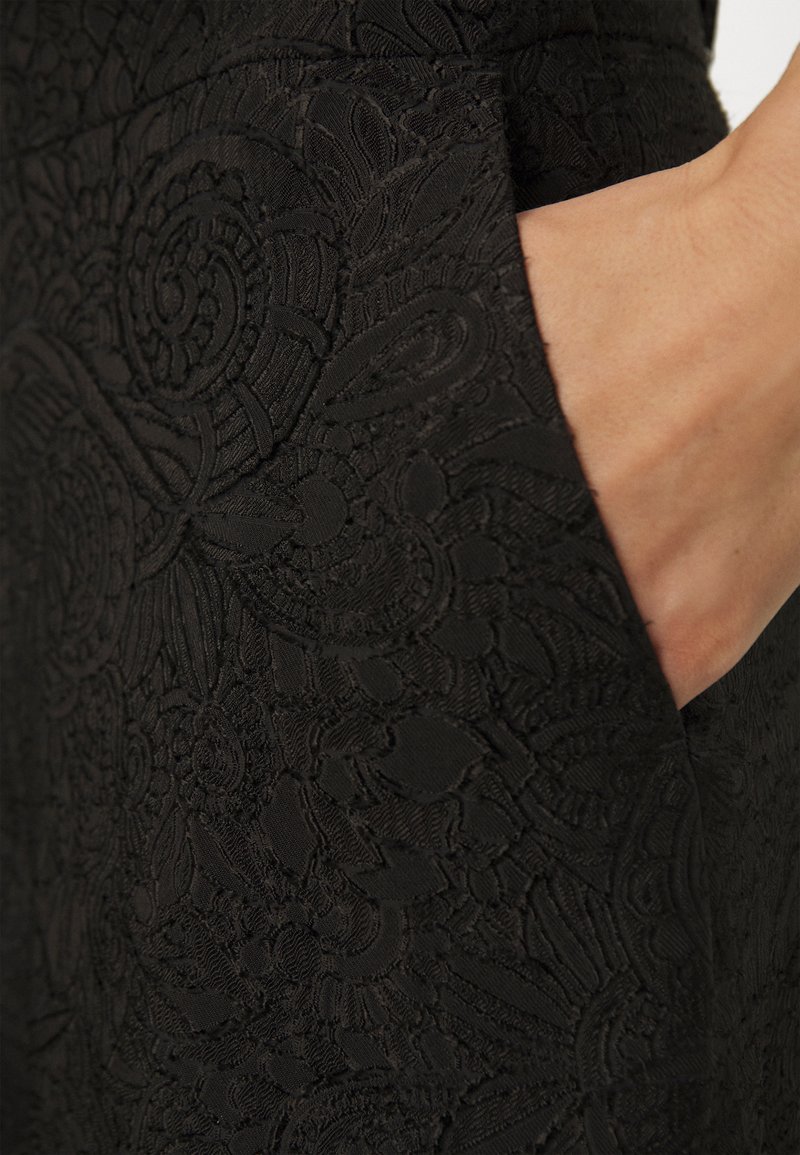 Schwarzes Spitzenkleid mit aufwendigen Blumenmustern, das eine Seitentasche und strukturierten Stoff aufweist. Eine Nahaufnahme hebt die detaillierte Stickerei hervor.