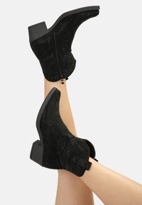 Botines de ante negros con cierre de cremallera, costuras decorativas y un tacón grueso. Superficie texturizada con pequeños embellecimientos.