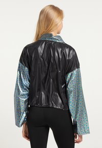 Sort og iriserende jakke med en skinnende sort krop og mønstrede metalærmer. Har en krave og elastisk kant.