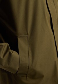 Olive green jacket featuring a smooth texture, angled pocket, and stitched accents. The fabric appears lightweight and durable.