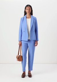 Light blue blazer and matching trousers in a tailored fit, paired with a pleated white top and brown leather handbag. Flat shoes with animal print.