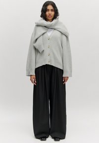 Helles graues Oversized-Cardigan mit großen Knöpfen und gerippten Bündchen, über einem weißen Oberteil getragen. Kombiniert mit weiten schwarzen Hosen.