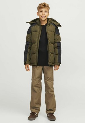 Veste matelassée en olive avec des accents noirs, fermeture éclair à l'avant et capuche amovible, associée à un pantalon marron clair et des baskets marron.