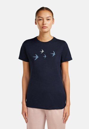 Woman wearing a navy blue t-shirt with four flying bird graphics across the chest, paired with light pink pants, standing against white background.