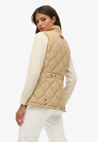 Gilet matelassé beige avec un col haut, doté d'une taille ceinturée et de boutons-pression. Porté par-dessus un haut à manches longues blanc.