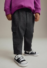 Graue Fischgrätenhose mit roten Streifen, ausgestattet mit einer Seitentasche. Kombiniert mit schwarzen Sneakers mit weißen Sohlen und Schnürsenkeln, getragen mit einem lila Pullover.