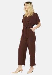 De bruine jumpsuit heeft een wikkelontwerp, korte mouwen, een tailleband en zijzakken, gemaakt van een gladde, lichte stof.