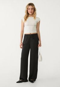 Schwarze weit geschnittene Hose mit elastischem Bund und Kordelzug, kombiniert mit einem enganliegenden weißen Croptop. Accessoires sind eine kleine weiße Tasche und schwarze Schuhe.