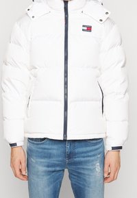 Hvid dunjakke med navy lynlåslukning, sidelommer og et logopatch på brystet. Har aftagelig hætte og elastiske manchetter.