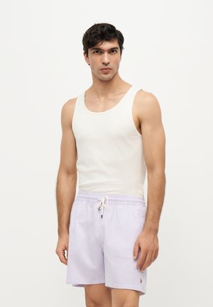 Jeune homme aux cheveux foncés portant un débardeur blanc et un short rayé violet clair avec cordon, debout devant un fond uni.