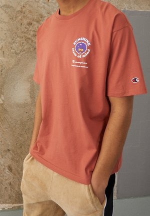 Person trägt ein rostfarbenes Champion-T-Shirt mit dem Text "Sunshine state of mind" und Grafik, Hände in den Taschen einer beigefarbenen Cordhose.