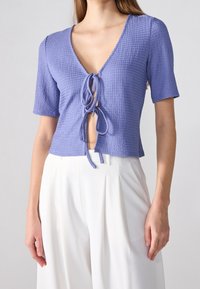 Levandulový top s krátkým rukávem s texturovaným povrchem a hlubokým výstřihem do V. Má zavazování vpředu a krátký střih.