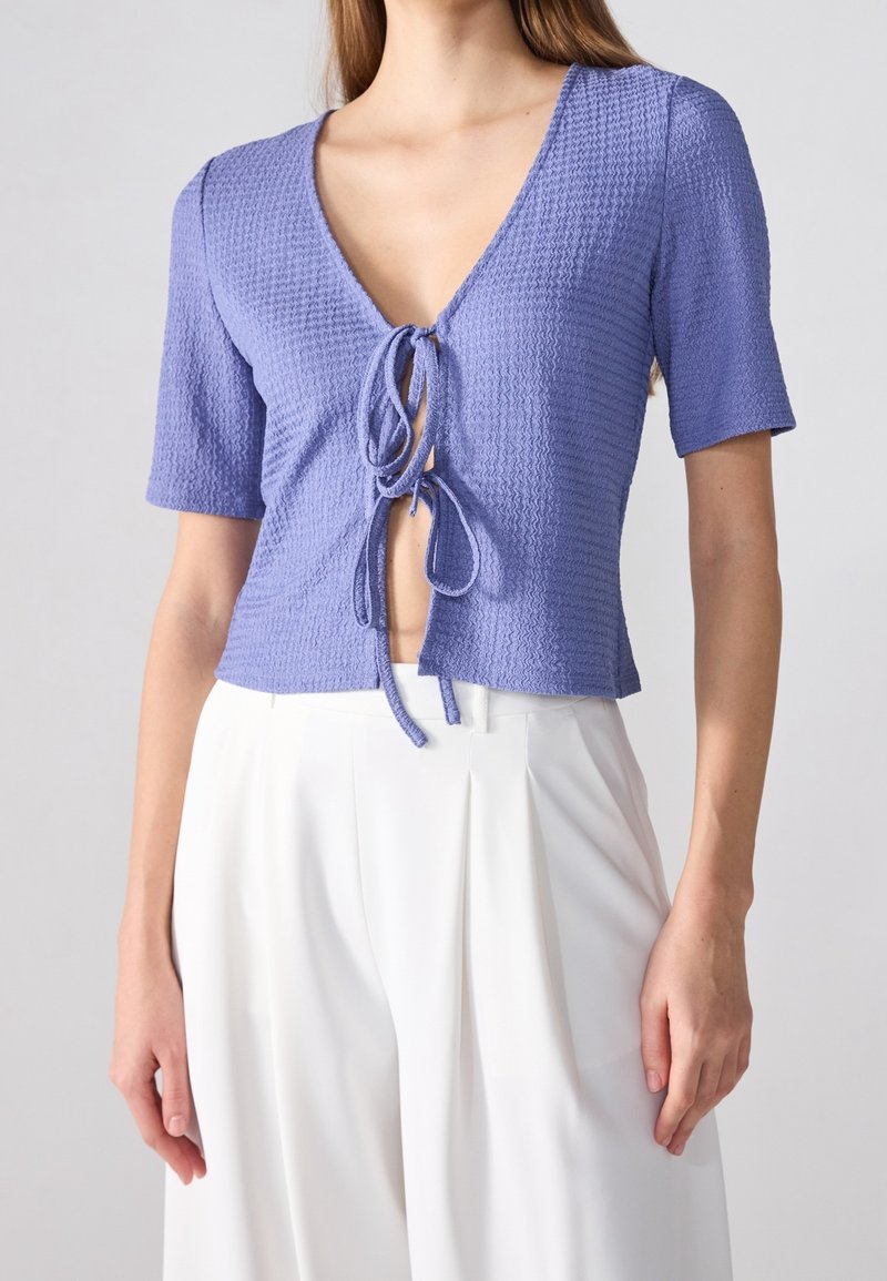 Levandulový top s krátkým rukávem s texturovaným povrchem a hlubokým výstřihem do V. Má zavazování vpředu a krátký střih.