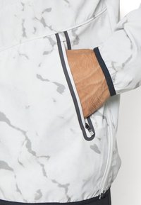 Main insérée dans la poche zippée d'une veste blanche à motif marbré avec un liseré noir sur la poche et le poignet.