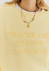 Žltá mikina s plyšovými textúrovanými nápismi "SNACKS ARE MY LOVE LANGUAGE." Viacfarebný perlový náhrdelník so zlatým príveskom.