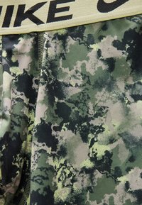 Κοντινό πλάνο υφάσματος με πράσινο και μαύρο σχέδιο παραλλαγής και κίτρινη μέση που φέρει το μερικό μαύρο κείμενο "NIKE."