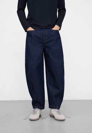 Person trägt dunkelblaue, lockere Jeans mit den Händen in den Taschen, kombiniert mit weißen Turnschuhen und einem schwarzen Langarmshirt, steht auf grauem Boden.