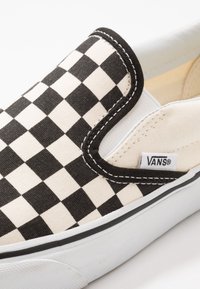 Zapato de lona slip-on en blanco y negro con diseño de tablero de ajedrez, con un collar acolchado, suela de goma blanca y costuras en contraste. Etiqueta de la marca visible.