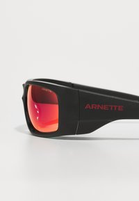 Lunettes de soleil noires avec des verres miroités rouges et une finition mate. Pr�ésente le logo "ARNETTE" en rouge sur le côté de la monture.
