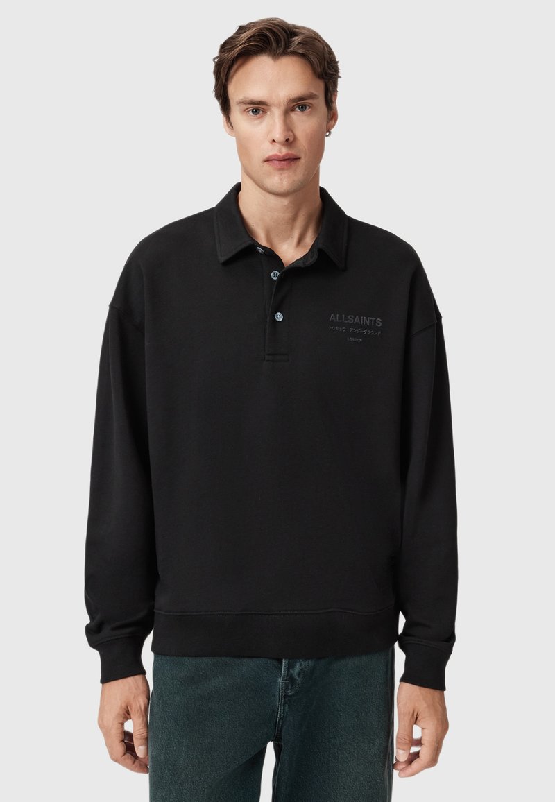 Mandlig model iført sort langærmet polo med "ALLSAINTS" logo og mørke jeans, stående mod en ensfarvet lys grå baggrund.