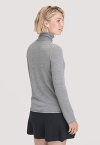 Blonde Frau in einem grauen Langarm-Pullover mit Rollkragen und einem schwarzen Strickrock, die seitlich vor einem einfarbigen Hintergrund steht.