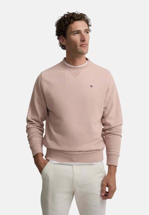 Hombre con el pelo rizado que lleva una sudadera rosa claro y pantalones blancos, mirando de lado con las manos en los bolsillos frente a un fondo liso.