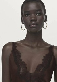 Bruine cami met kanten afwerking en een diep V-hals ontwerp. Model heeft kort haar en gouden oorringen. Gladde textuur, minimalistische stijl.