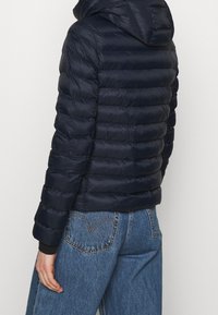 Giacca imbottita blu navy con cappuccio e motivo trapuntato orizzontale, caratterizzata da un design aderente e polsini elasticizzati. Indossata sopra jeans di denim blu.