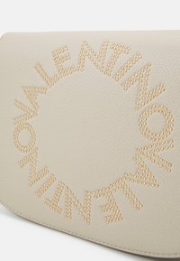 Borsa in pelle beige con una disposizione circolare di borchie dorate che formano la parola "Valentino". Texture liscia con angoli arrotondati.